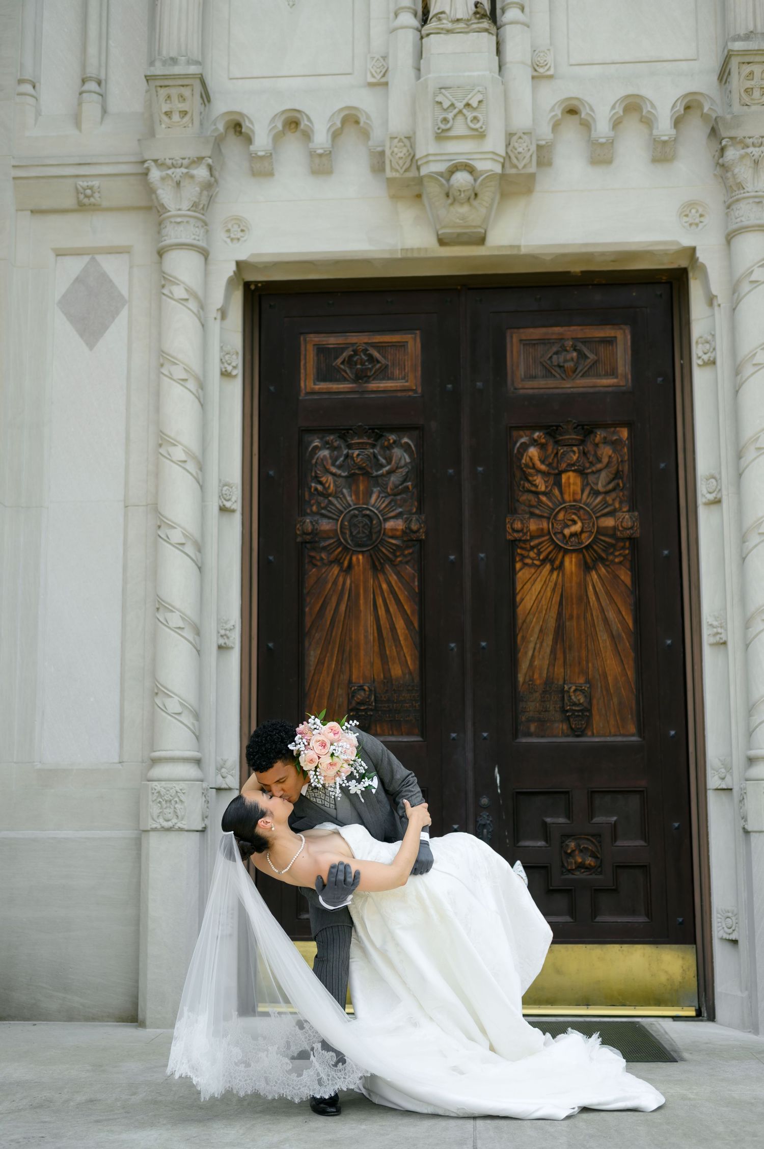 Bride and groom share a romantic dip kiss in front of ornate wooden doors of a white stone church.