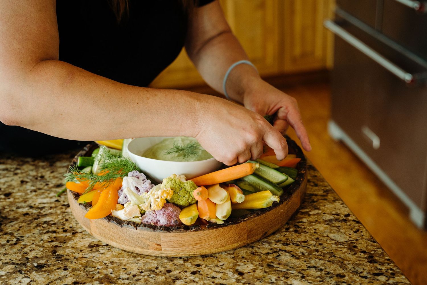 Woman arranging fresh vegetables and dip on a wooden charcuterie board on a granite countertop.