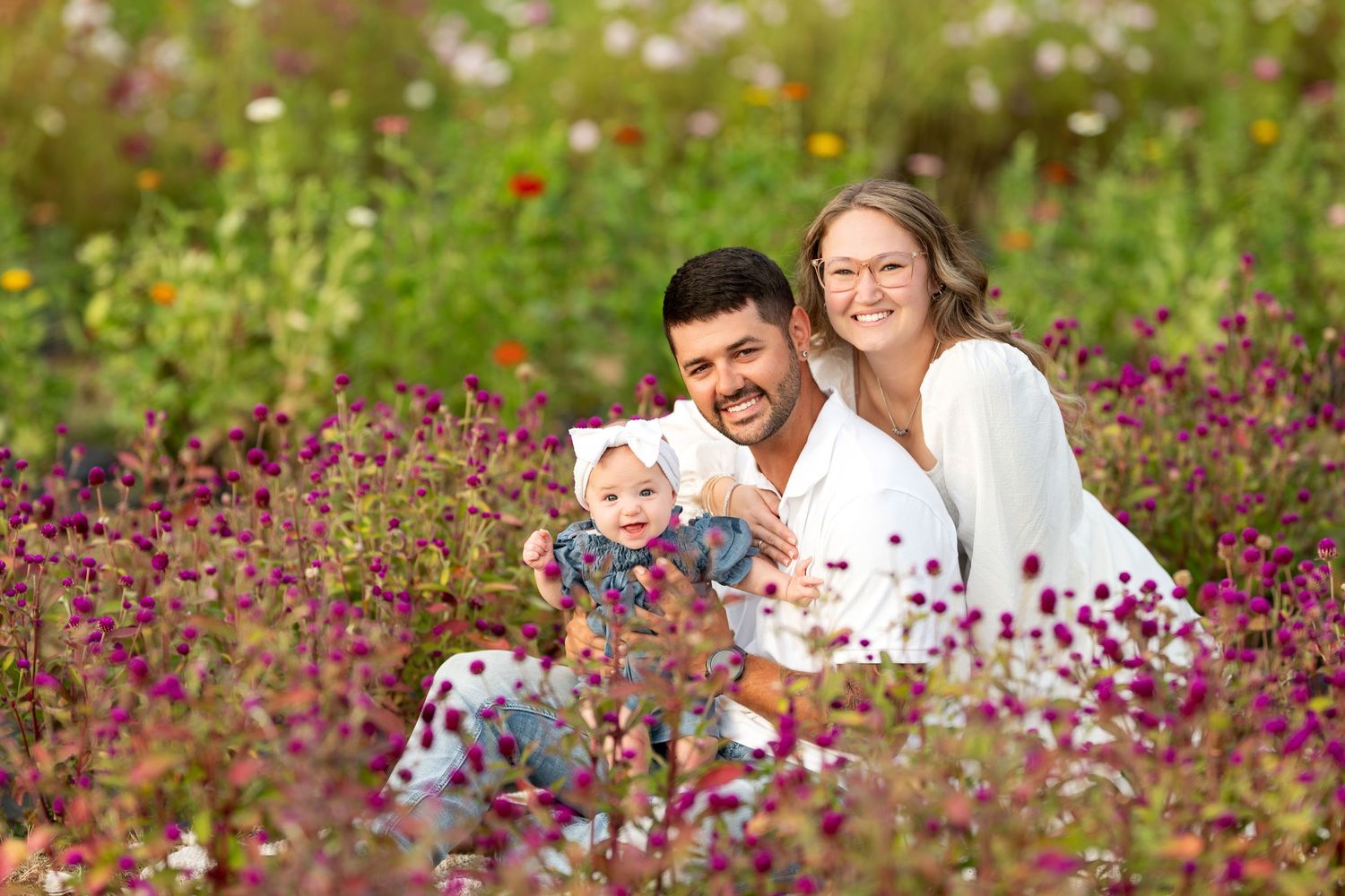 Happy family with baby sitting in a vibrant flower field, smiling couple holding infant among purple blooms outdoors.