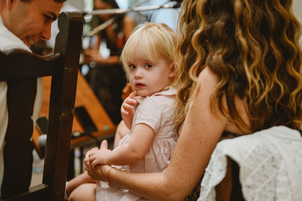 Adorable blonde toddler girl in pink dress sitting on lap during wedding ceremony.