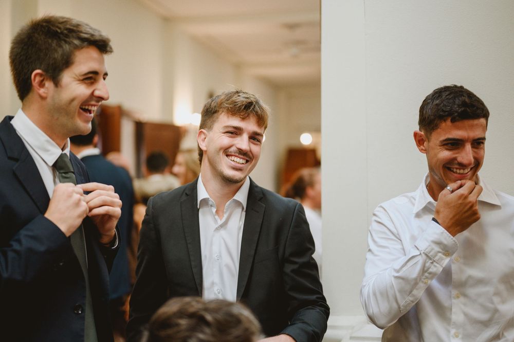Three young men in suits laughing and socializing at a formal wedding reception event.