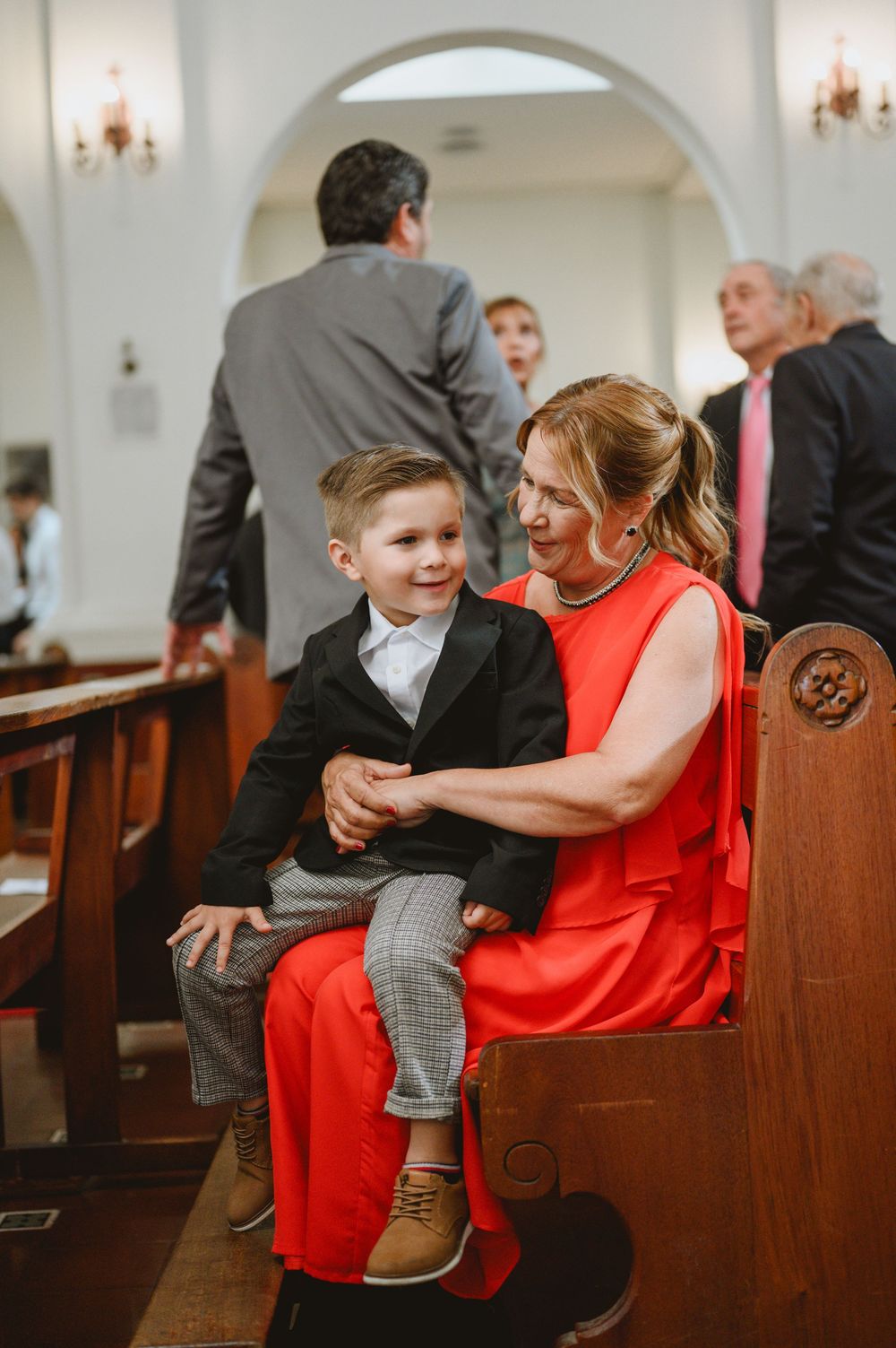 Woman in red dress sitting on church pew with young boy in suit during wedding ceremony.