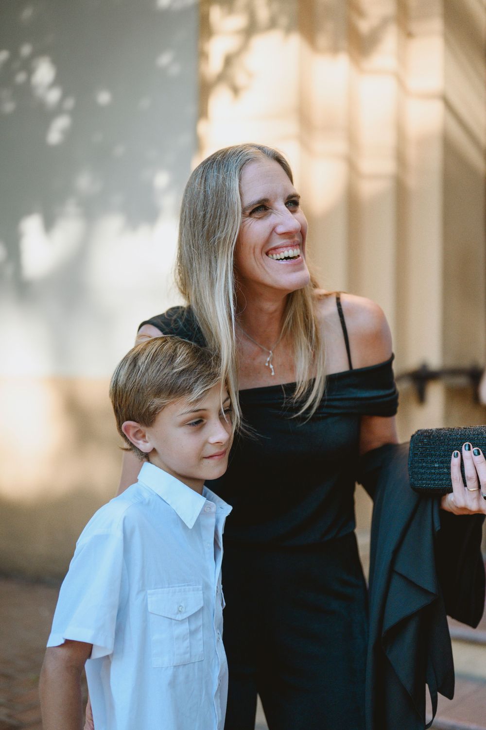 Smiling blonde woman in black off-shoulder dress posing with young boy at wedding venue.