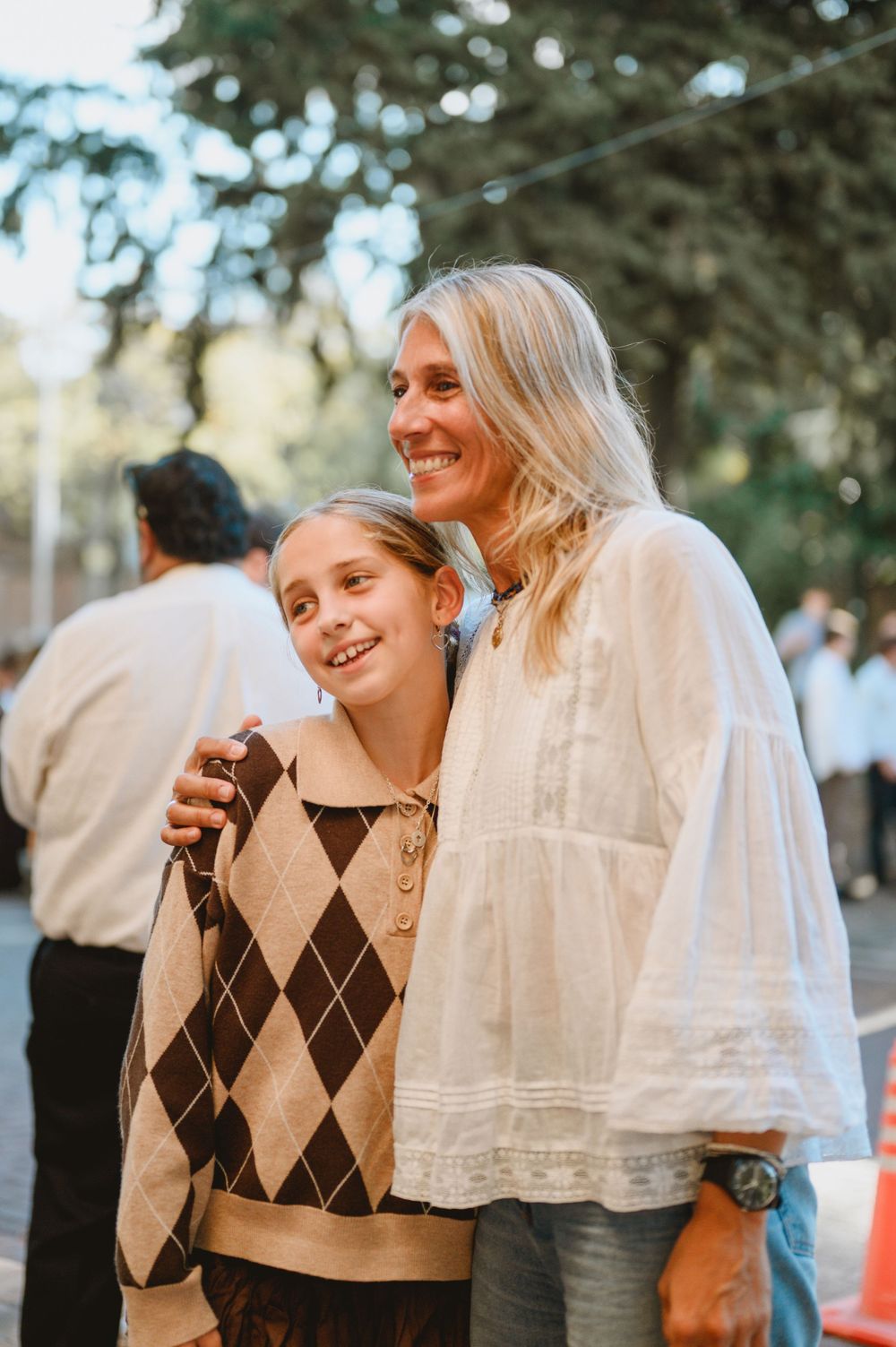 Blonde woman in white blouse posing with young girl in argyle sweater at outdoor wedding.