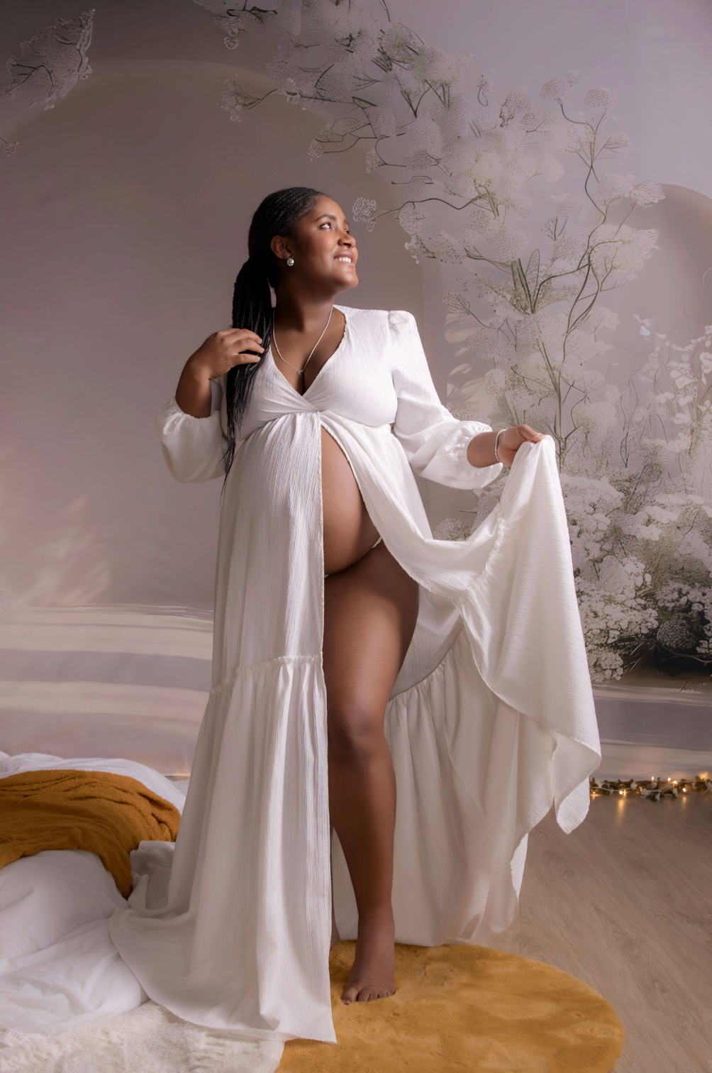 Pregnant woman in white flowing dress standing barefoot on a sandy beach at sunset.