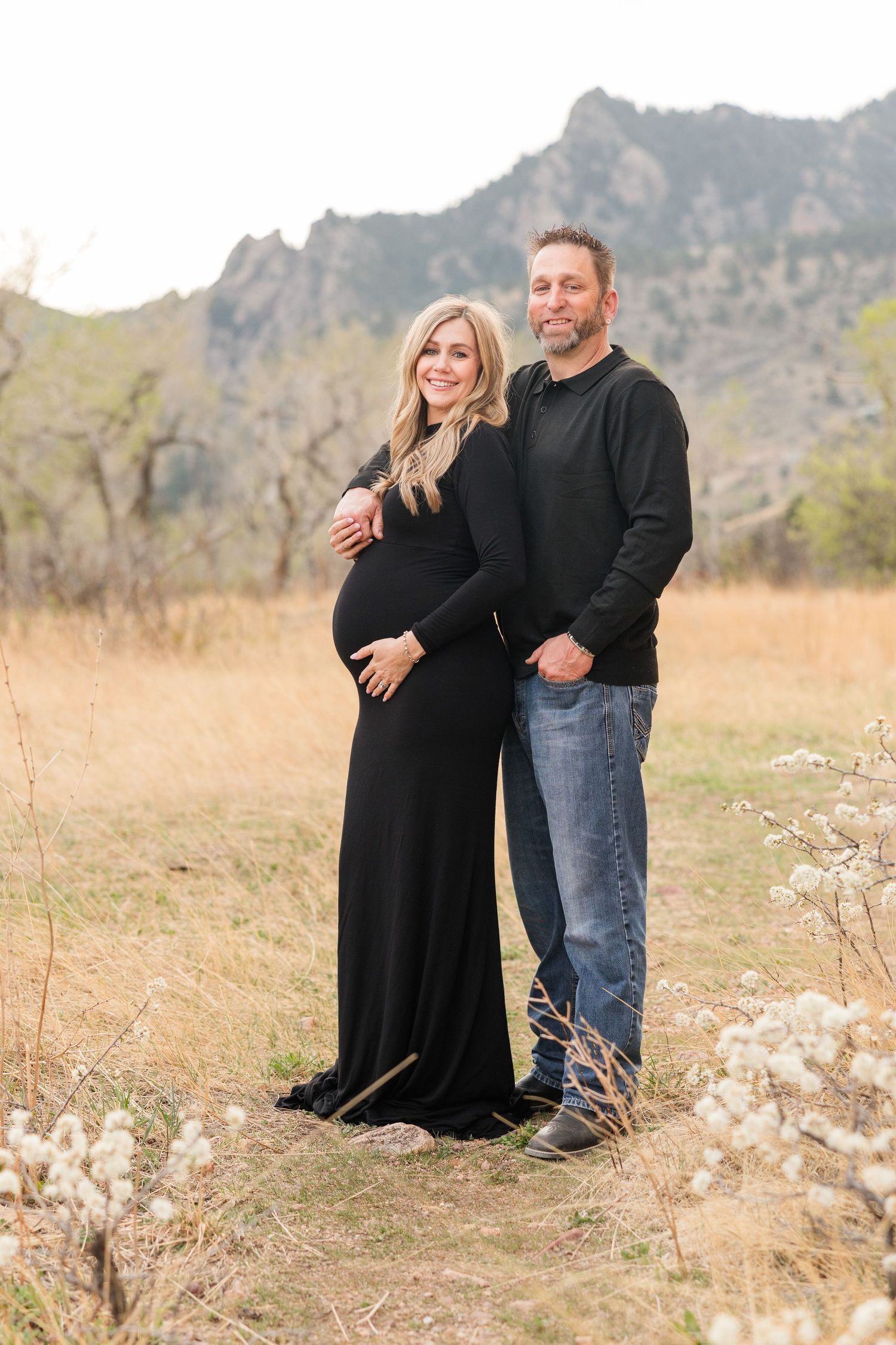 Pregnant couple posing outdoors in a mountain meadow, woman in black maternity dress, man in black sweater and jeans.