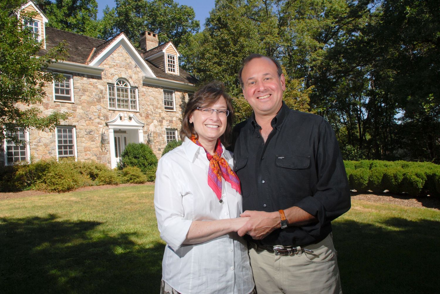 Couple smiling together in front of a stone colonial home surrounded by lush green trees.