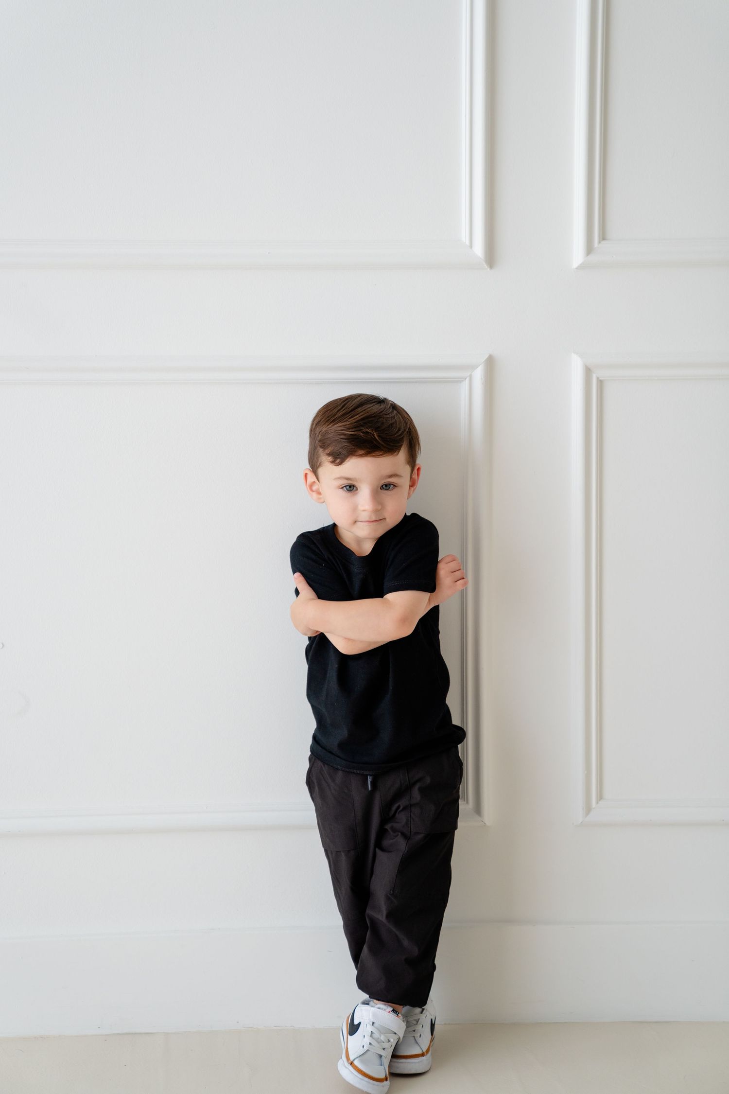 A young boy in a black t-shirt and pants stands against a white wall with arms crossed, wearing white sneakers.
