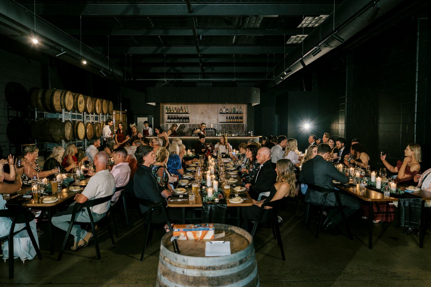 Guests dining at long tables in a rustic winery event space with wooden barrels and warm candlelight ambiance.