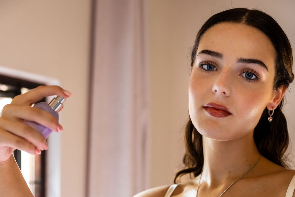 Young woman applying perfume spray, wearing elegant makeup with dark lips and a pearl earring, in a softly lit room.