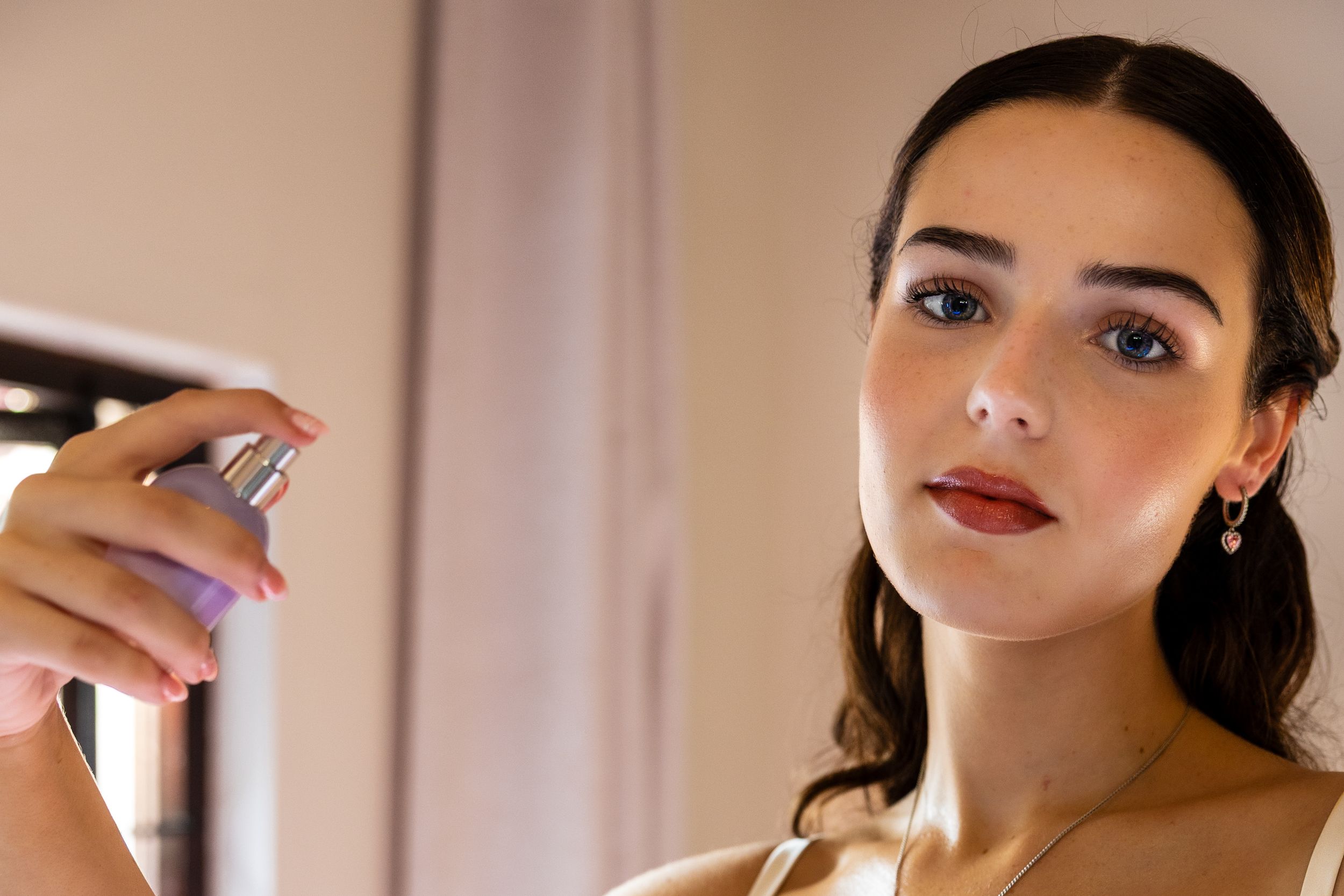 Young woman applying perfume spray, wearing elegant makeup with dark lips and a pearl earring, in a softly lit room.