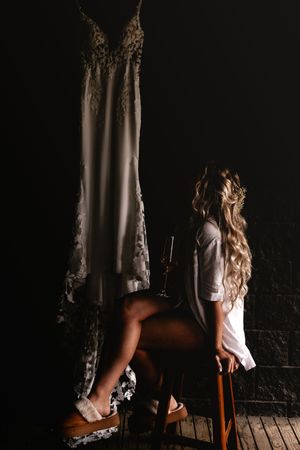 A bride sits on a chair gazing at her lace wedding dress hanging in a dark, moody room.