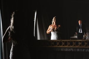 Bride in white gown stands in dramatic lighting at ornate mirror as her father sees in her wedding gown for the first time.