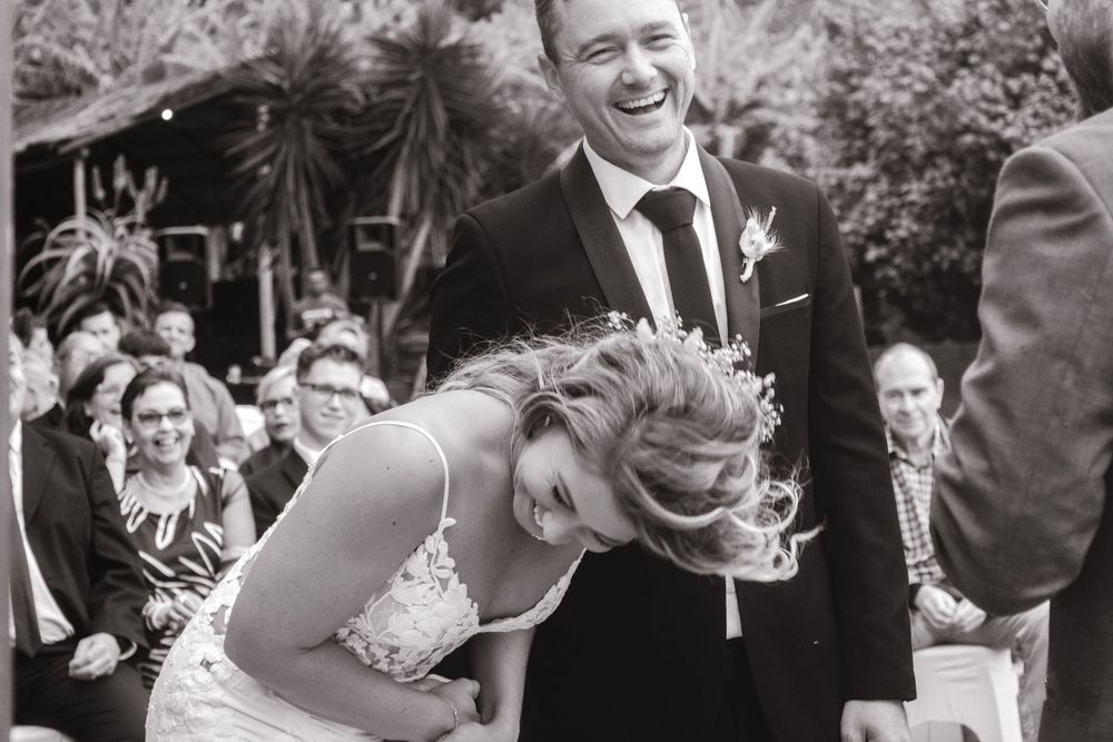 Black and white photograph of laughing bride and groom during outdoor wedding ceremony with guests seated behind them.