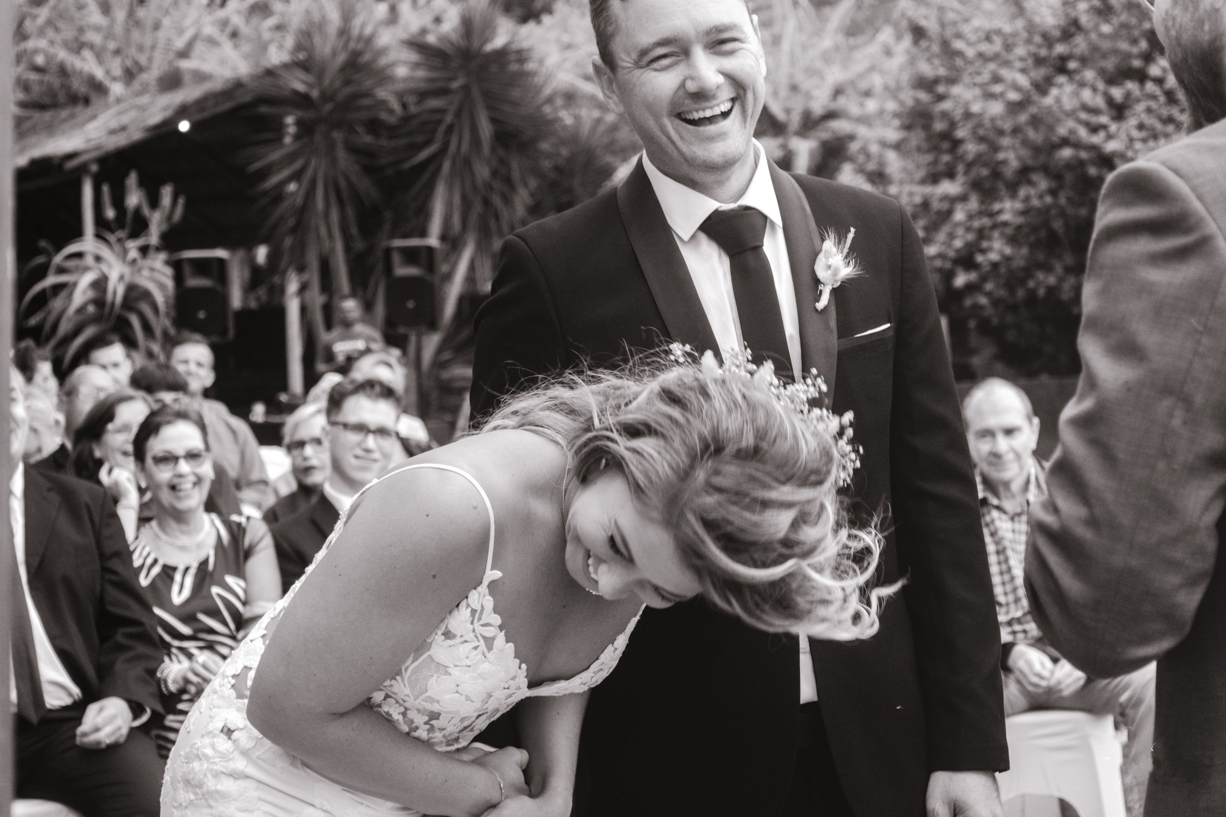 Black and white photograph of laughing bride and groom during outdoor wedding ceremony with guests seated behind them.