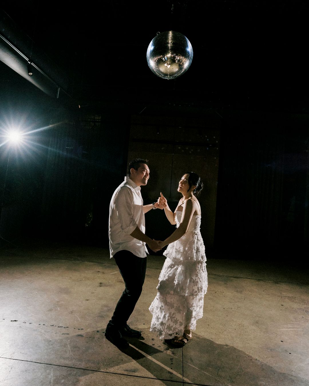 Couple dancing under a disco ball in a dimly lit venue, bride in ruffled white gown holding hands with groom in white shirt.