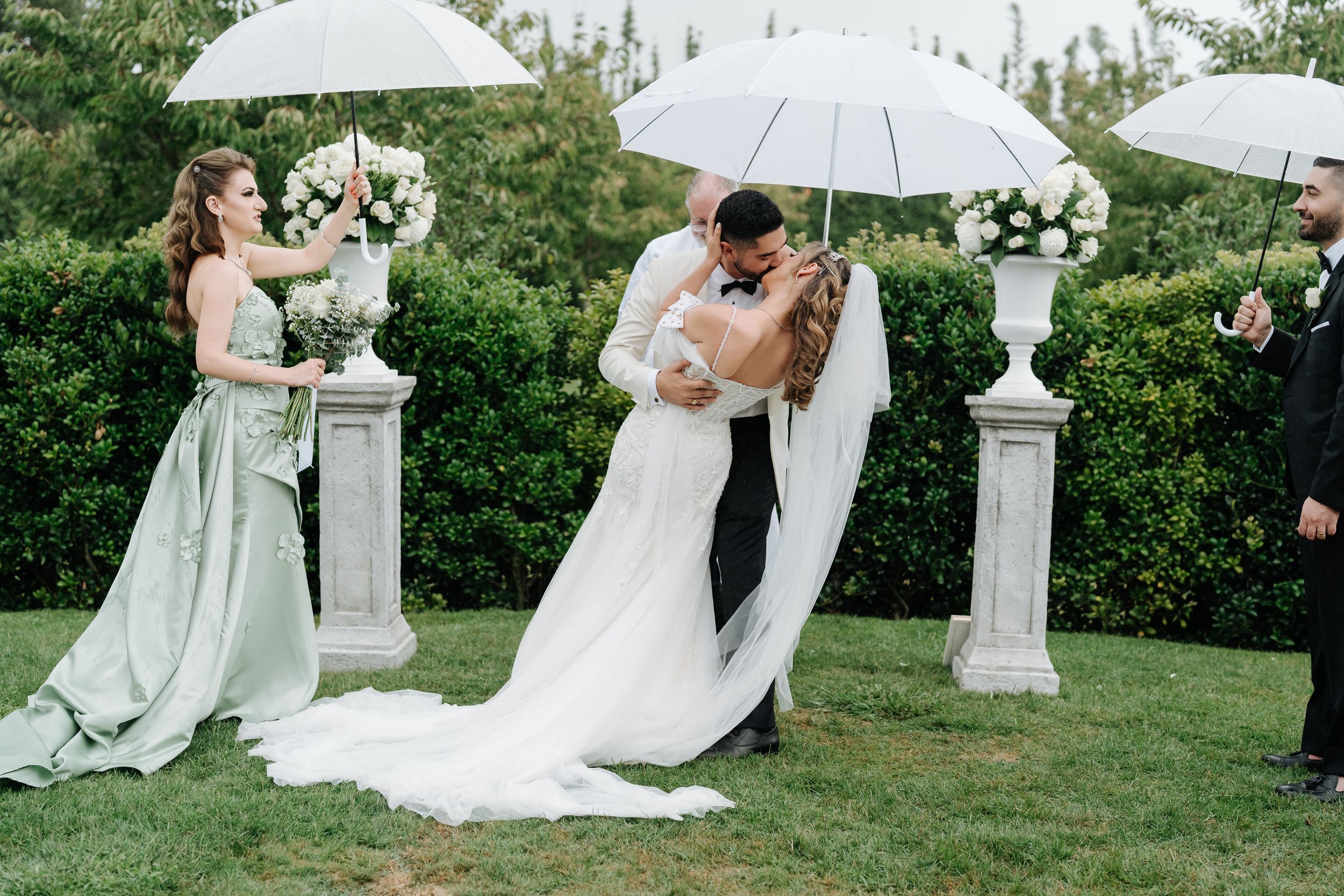 Bride and groom share a romantic dip kiss at outdoor wedding ceremony with white umbrellas and floral arrangements.
