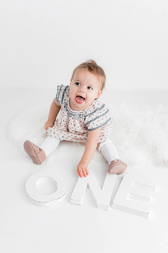 A happy baby girl in a floral dress sits on a white fur rug beside large white ONE letters for her first birthday photo.