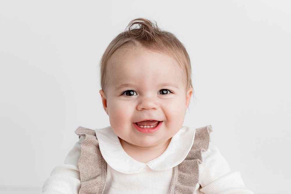 Smiling baby girl with tiny hair bun wearing white ruffle collar onesie and tan overalls against white background.
