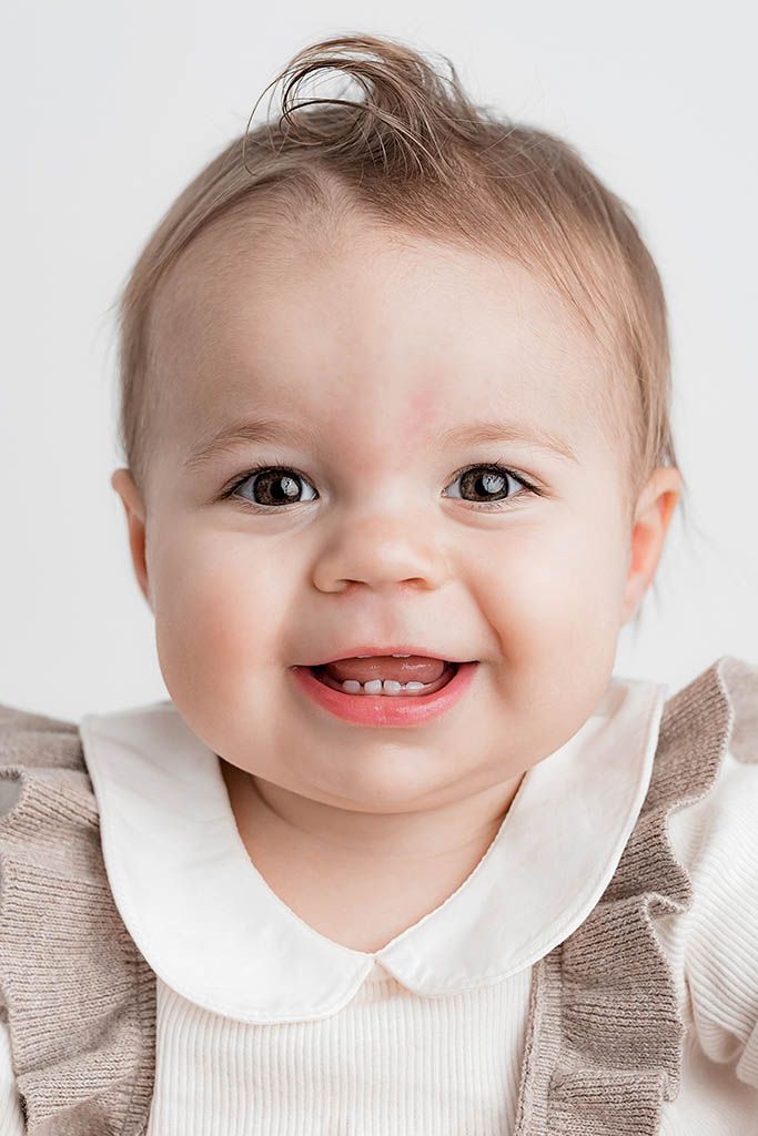 Smiling baby girl with brown eyes wearing a white collared shirt and beige ruffle outfit against a white background.