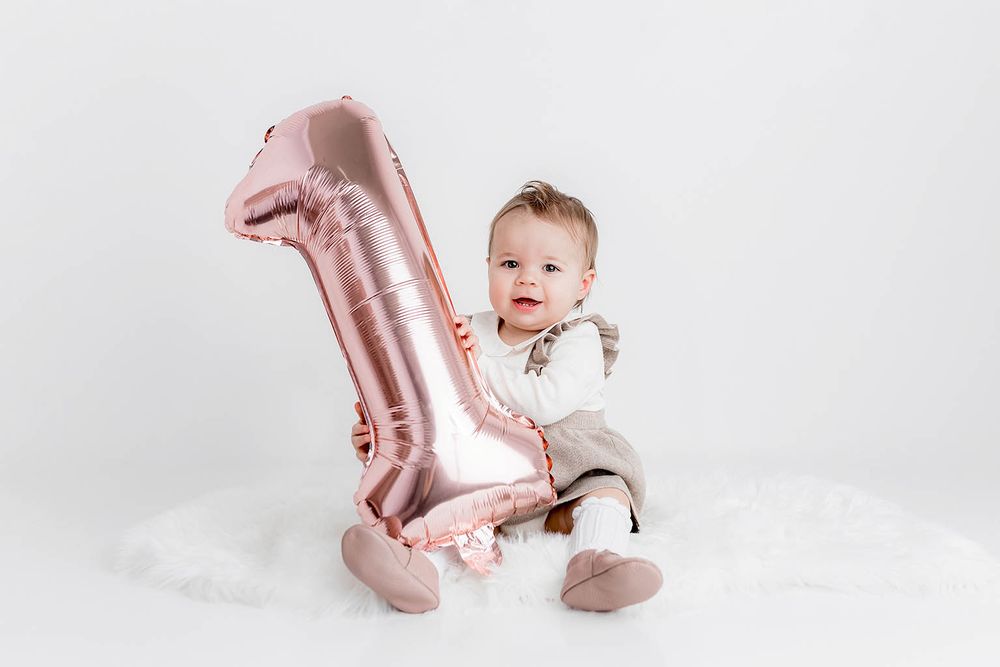 Adorable baby sitting beside a rose gold number one balloon celebrating first birthday on white background.