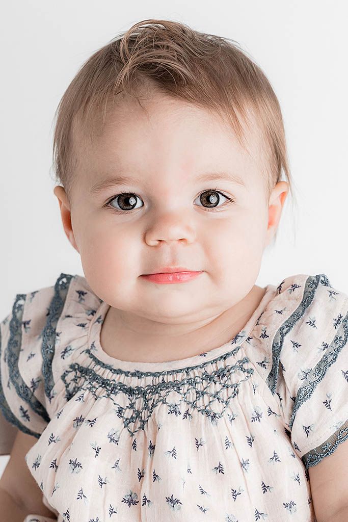 Close-up portrait of a baby girl with brown eyes wearing a smocked floral dress with blue lace trim.