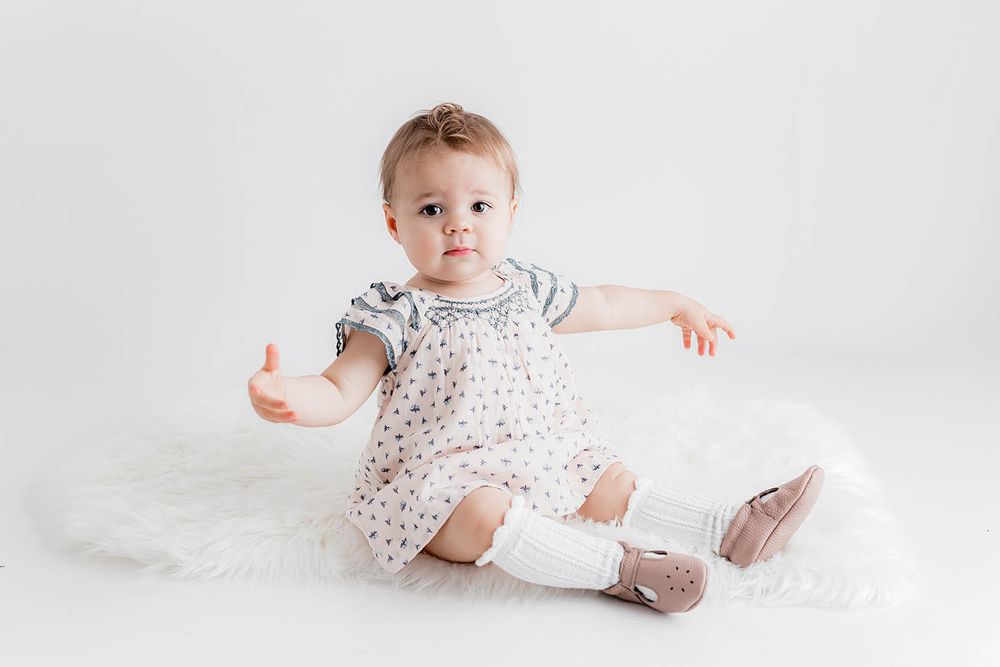 Baby girl in a floral smocked dress and white socks sitting on a fluffy white rug, reaching out her arm.