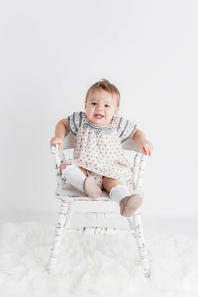 Happy baby girl in floral smocked dress sitting in vintage white wooden chair on fluffy white rug.