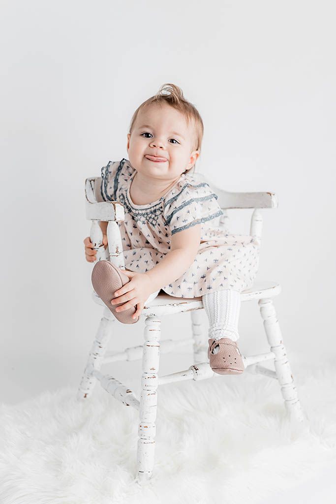 Smiling baby girl sitting on a white wooden chair in a light floral outfit with pink shoes on a fluffy white rug.