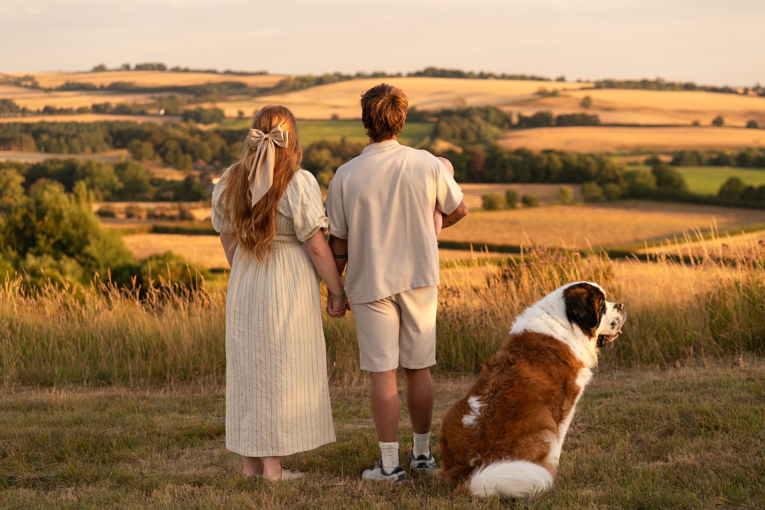 Couple and Saint Bernard dog enjoying golden hour sunset views over rolling green countryside fields.