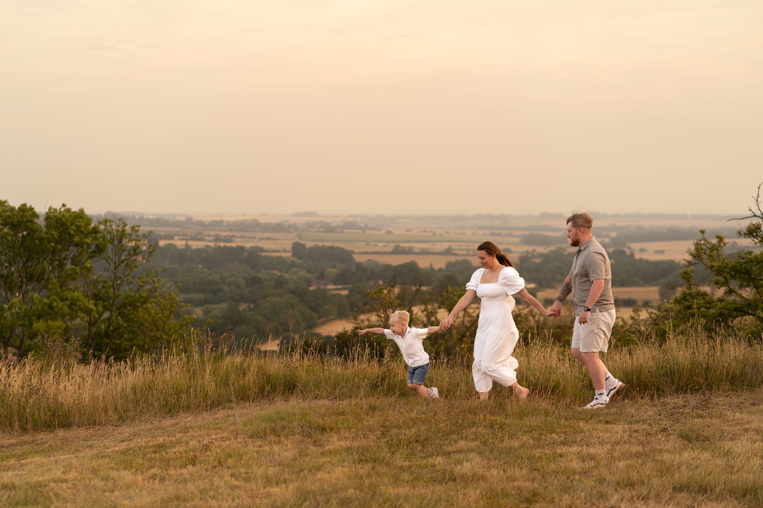 Family of three walking hand-in-hand on a grassy hilltop at golden hour with scenic countryside views.