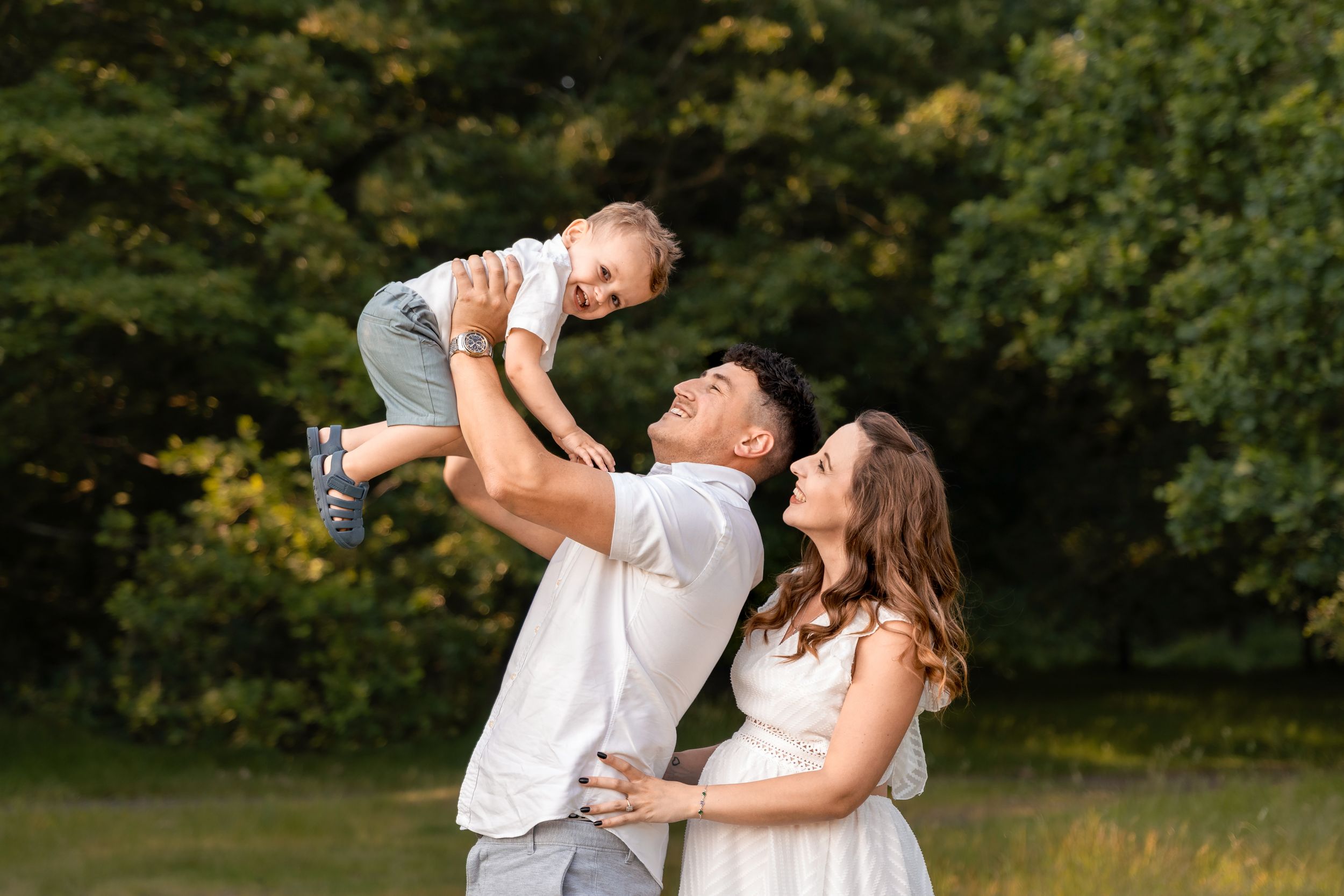 Happy family outdoors, father lifting toddler while mother watches, smiling together in a lush green park setting.