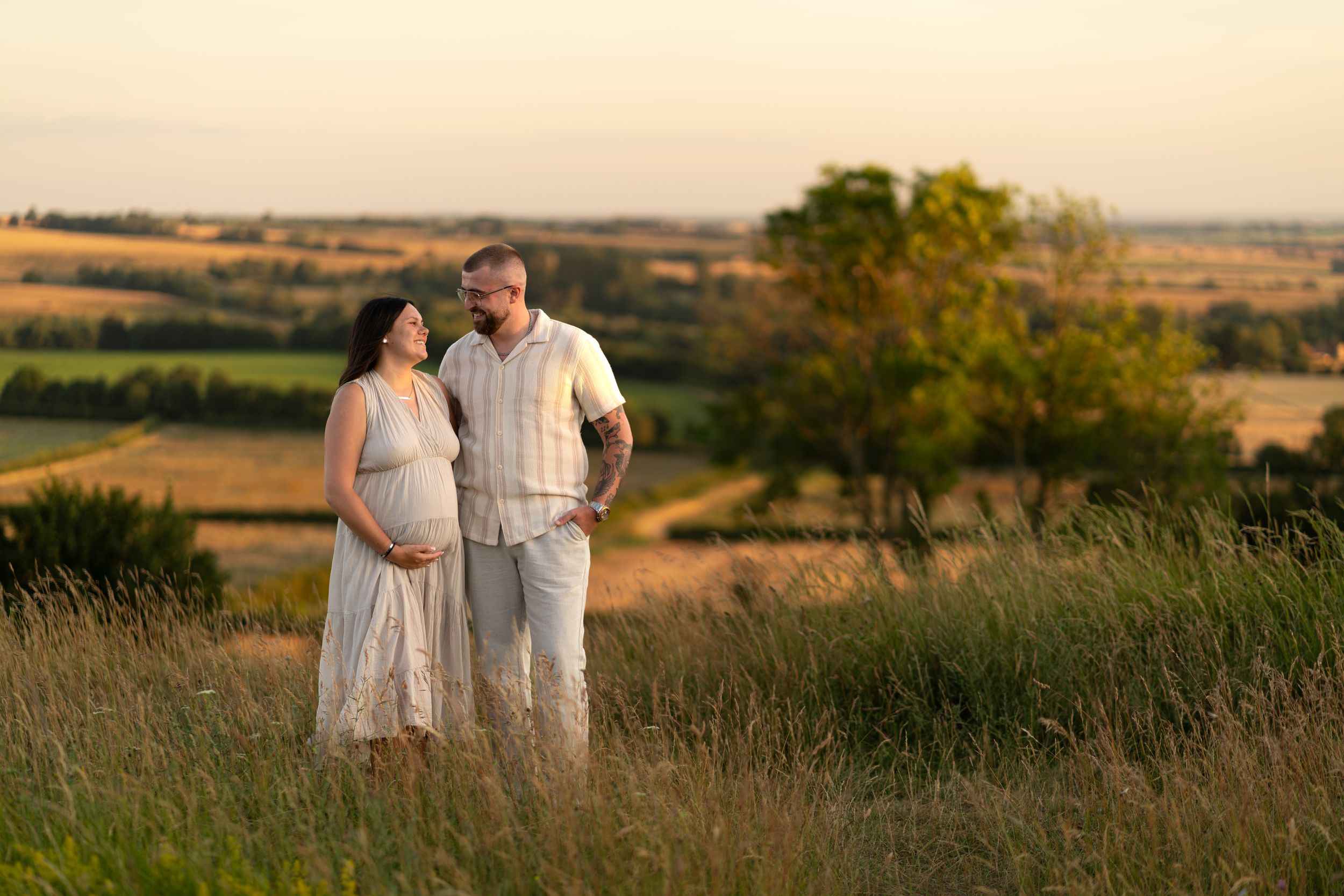 Pregnant couple standing in golden hour sunlight in a lush green field with scenic countryside views.