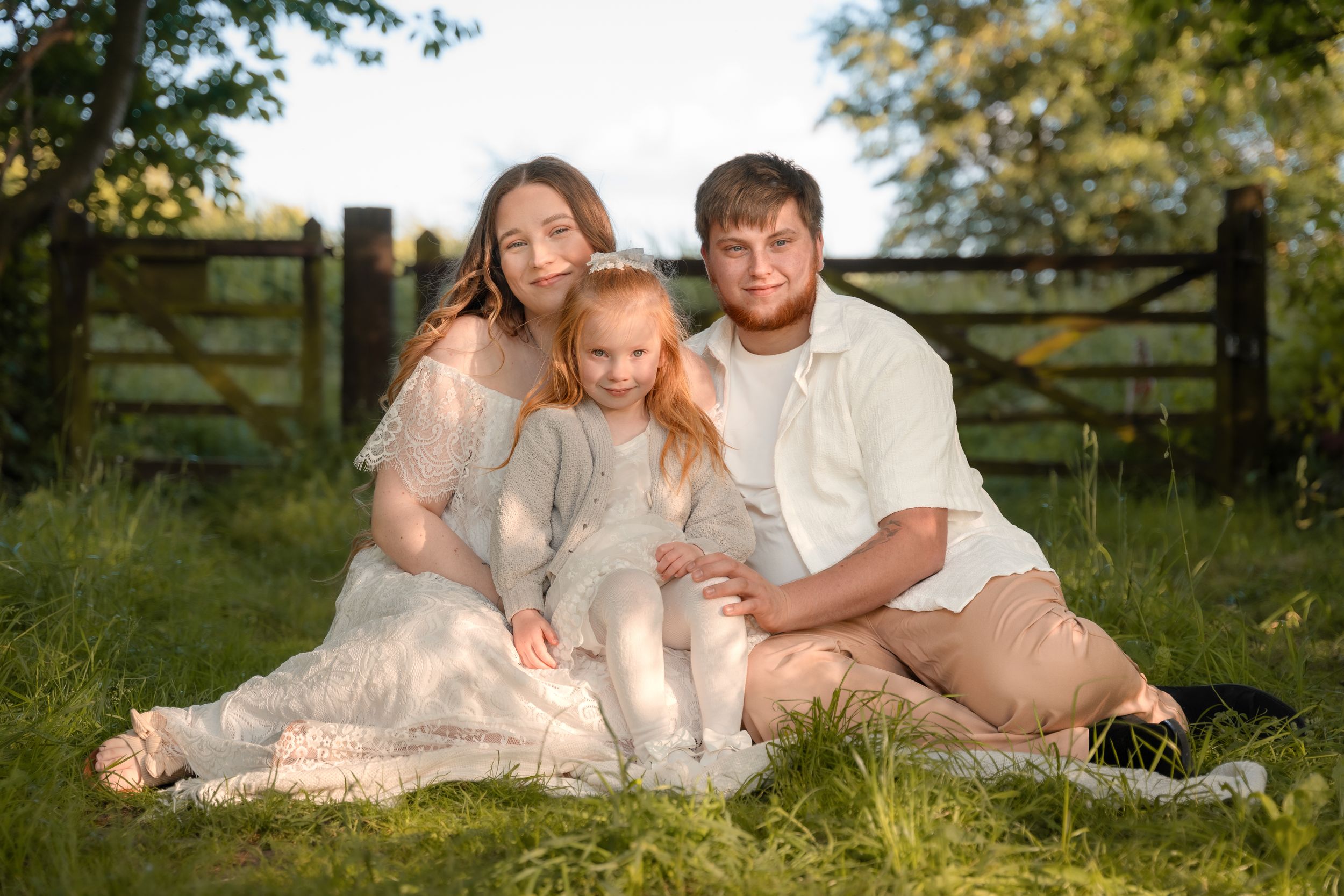 Family of three sitting on grass blanket outdoors, parents and young blonde girl posing near wooden fence in golden light.