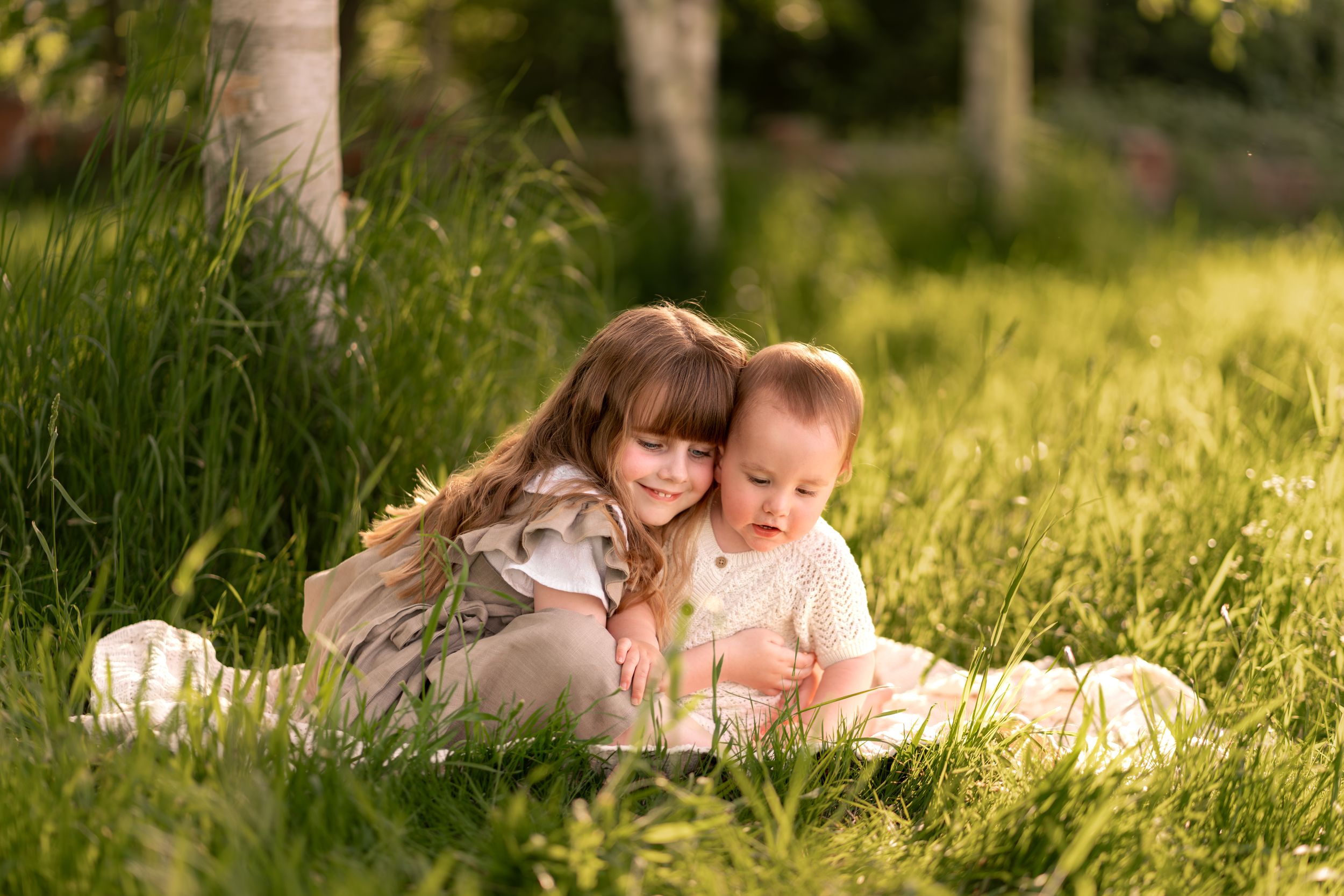 Two young children sitting in lush green grass outdoors, older girl hugging baby sibling in warm golden sunlight.
