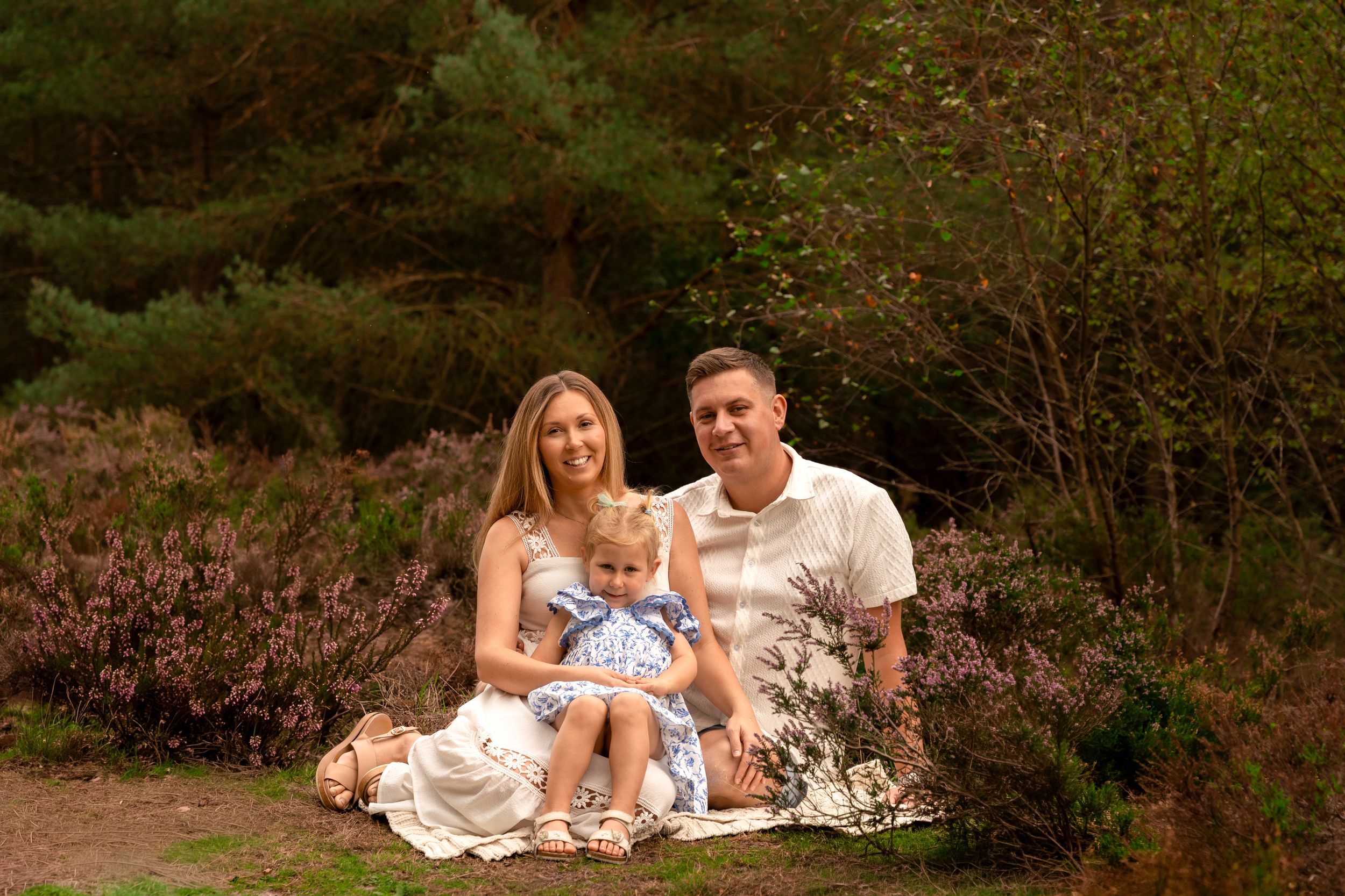 A happy family of three sits outdoors on a blanket surrounded by heather and pine trees in a scenic nature setting.