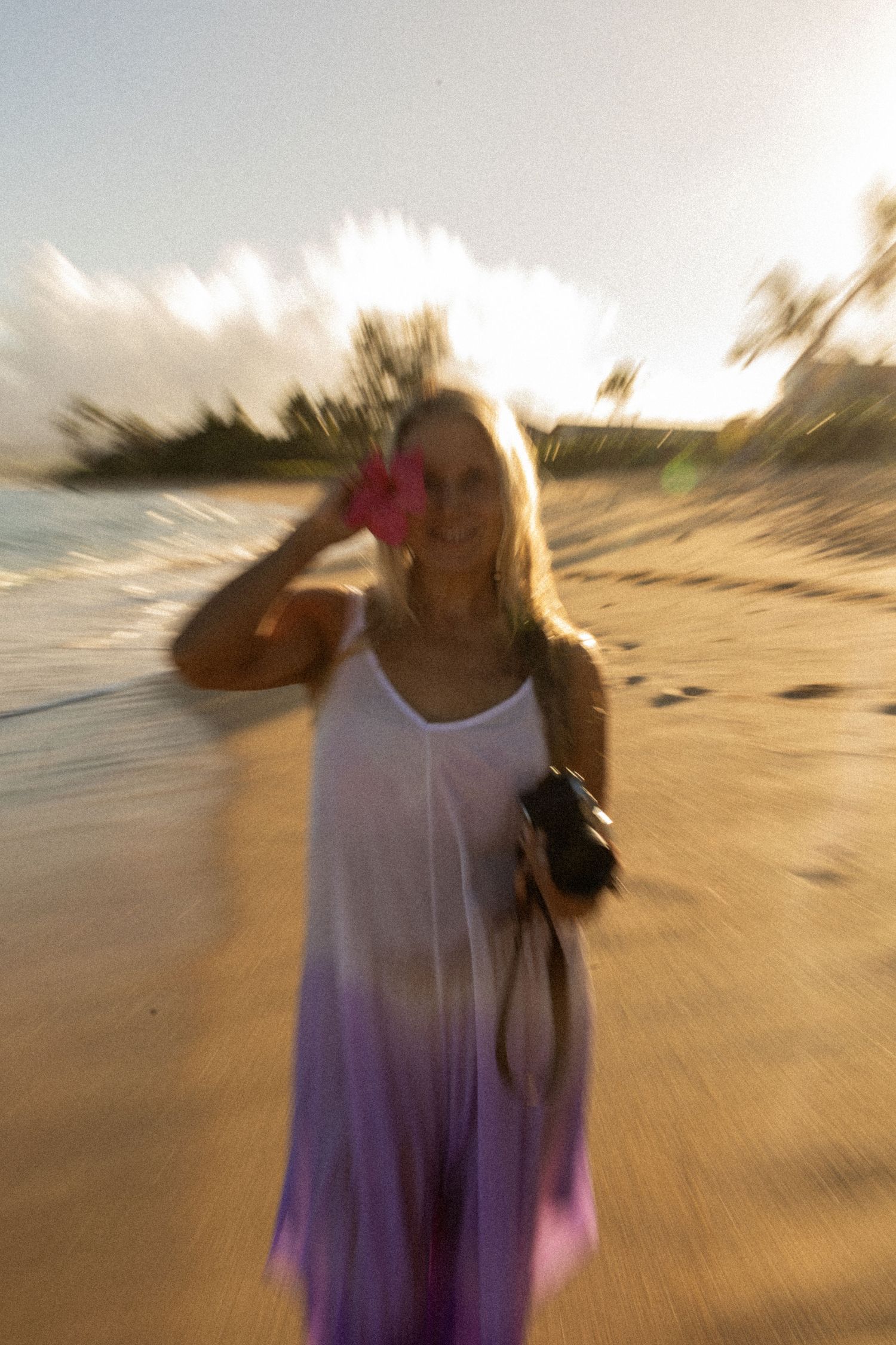 Blond woman in white and purple maxi dress holding camera on sunny tropical beach at golden hour on Oahu, Hawaii.