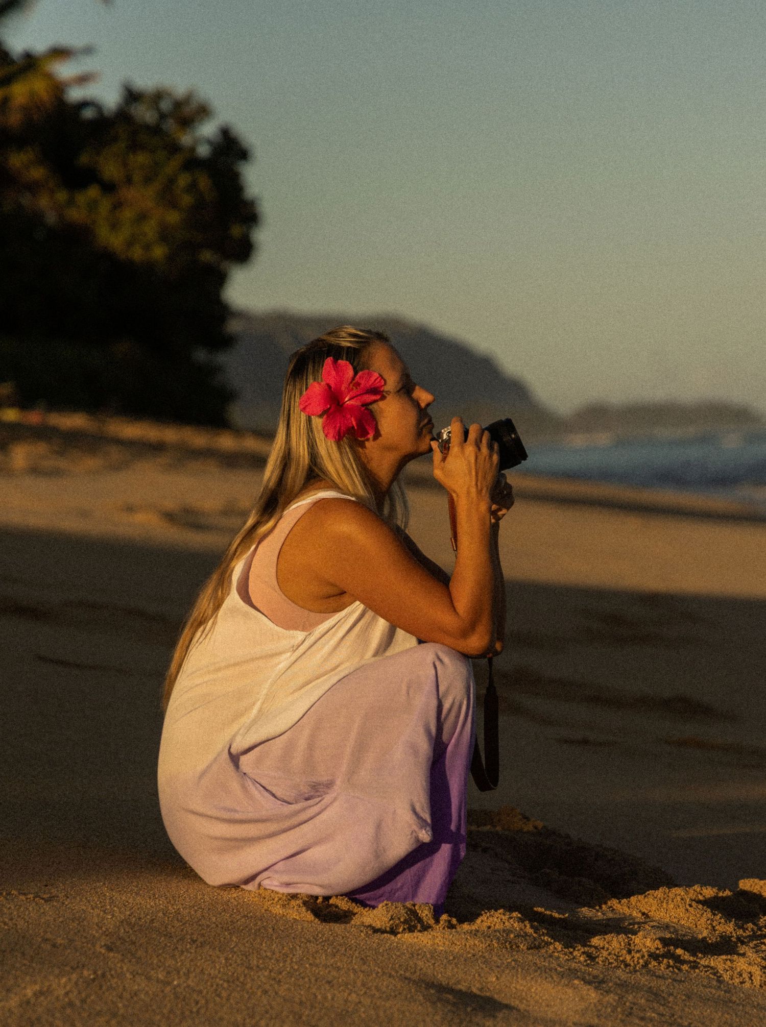Woman with pink flower in hair crouching on sandy beach photographing sunset with mountains in background on Oahu, Hawaii.