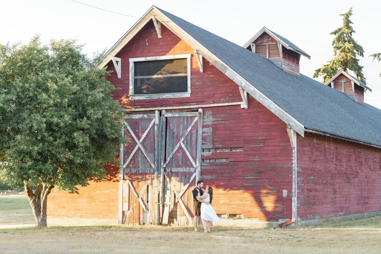 Couple embracing in front of a rustic red barn at golden hour on a scenic farm wedding venue.