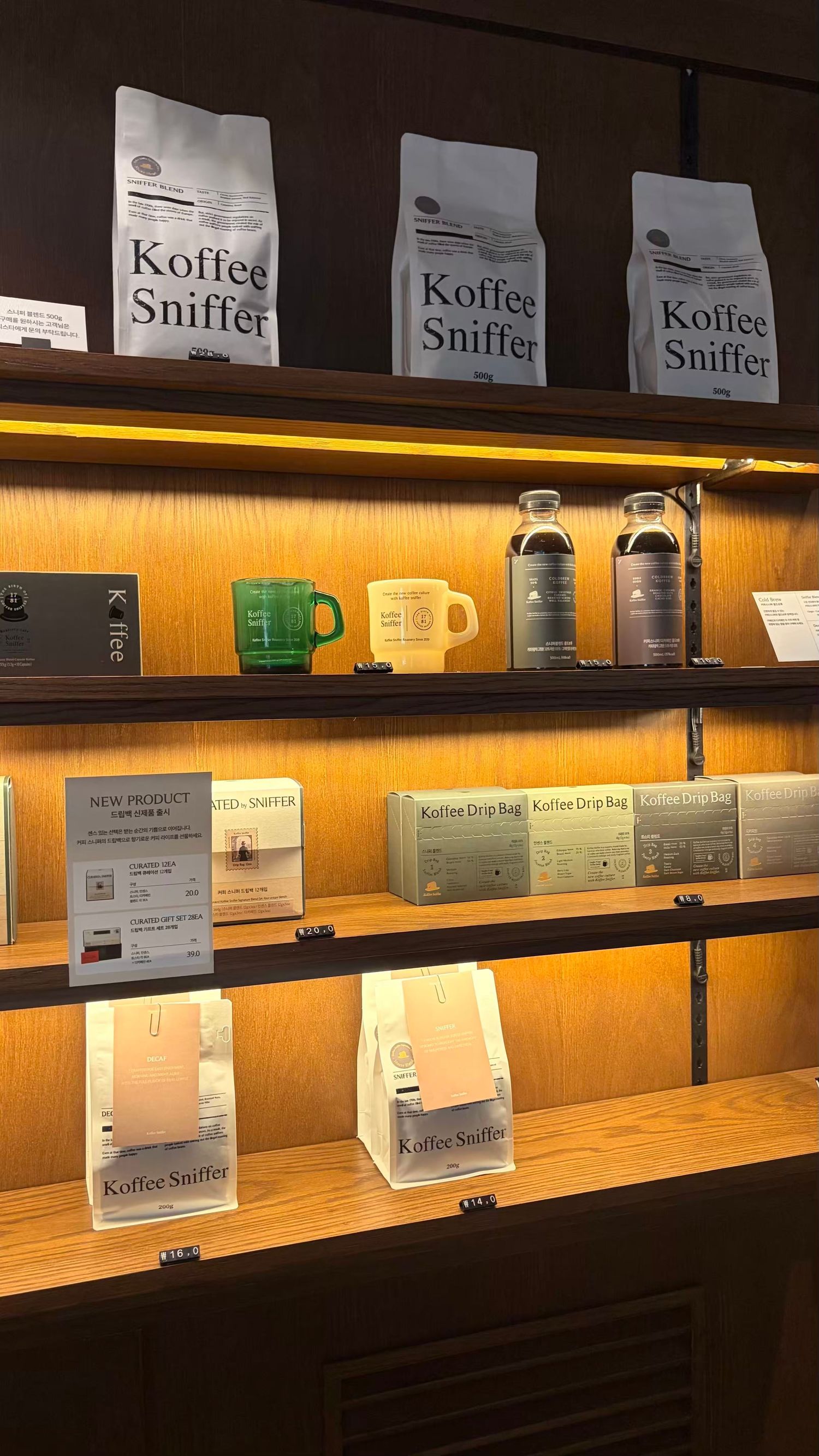 Koffee Sniffer coffee shop display shelf featuring coffee beans, drip bags, mugs, and cold brew bottles with warm lighting.