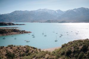 Scenic turquoise mountain lake with sailboats anchored near shoreline and snow-capped peaks beyond.