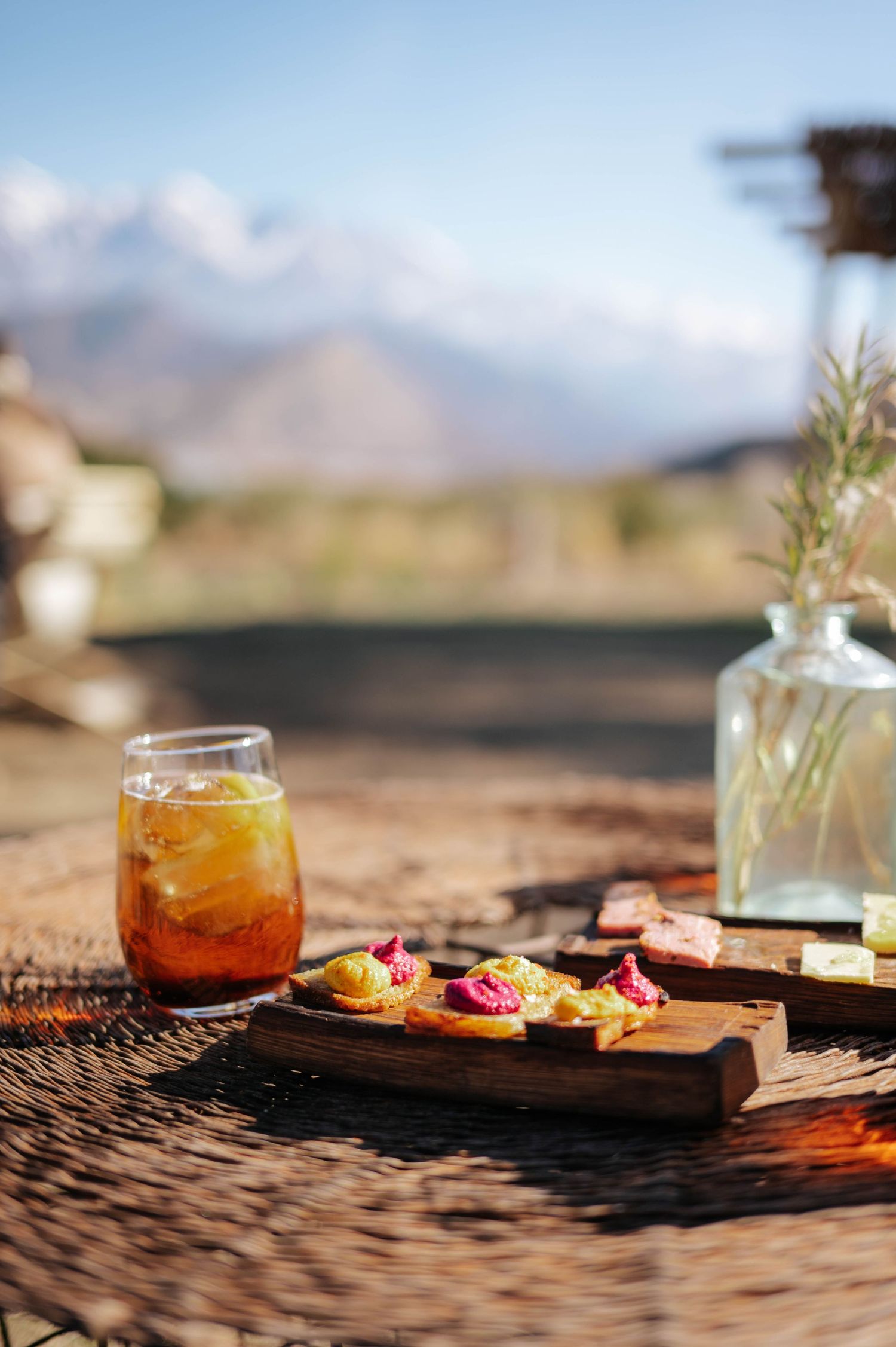 Iced cocktail and colorful appetizers on wooden board with mountain landscape in background.