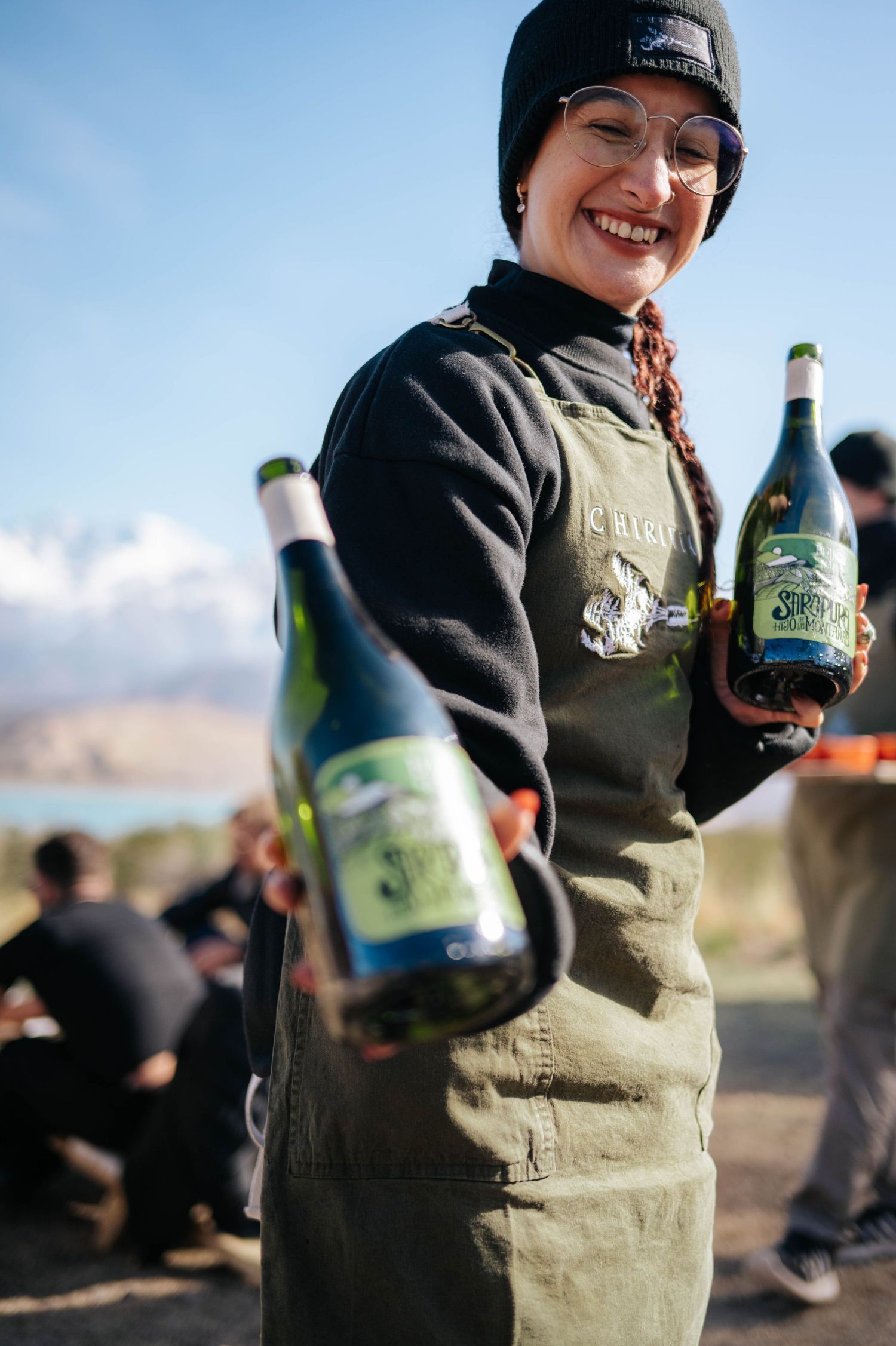 Smiling server holding green beer bottles wearing apron and beanie at outdoor mountain event.