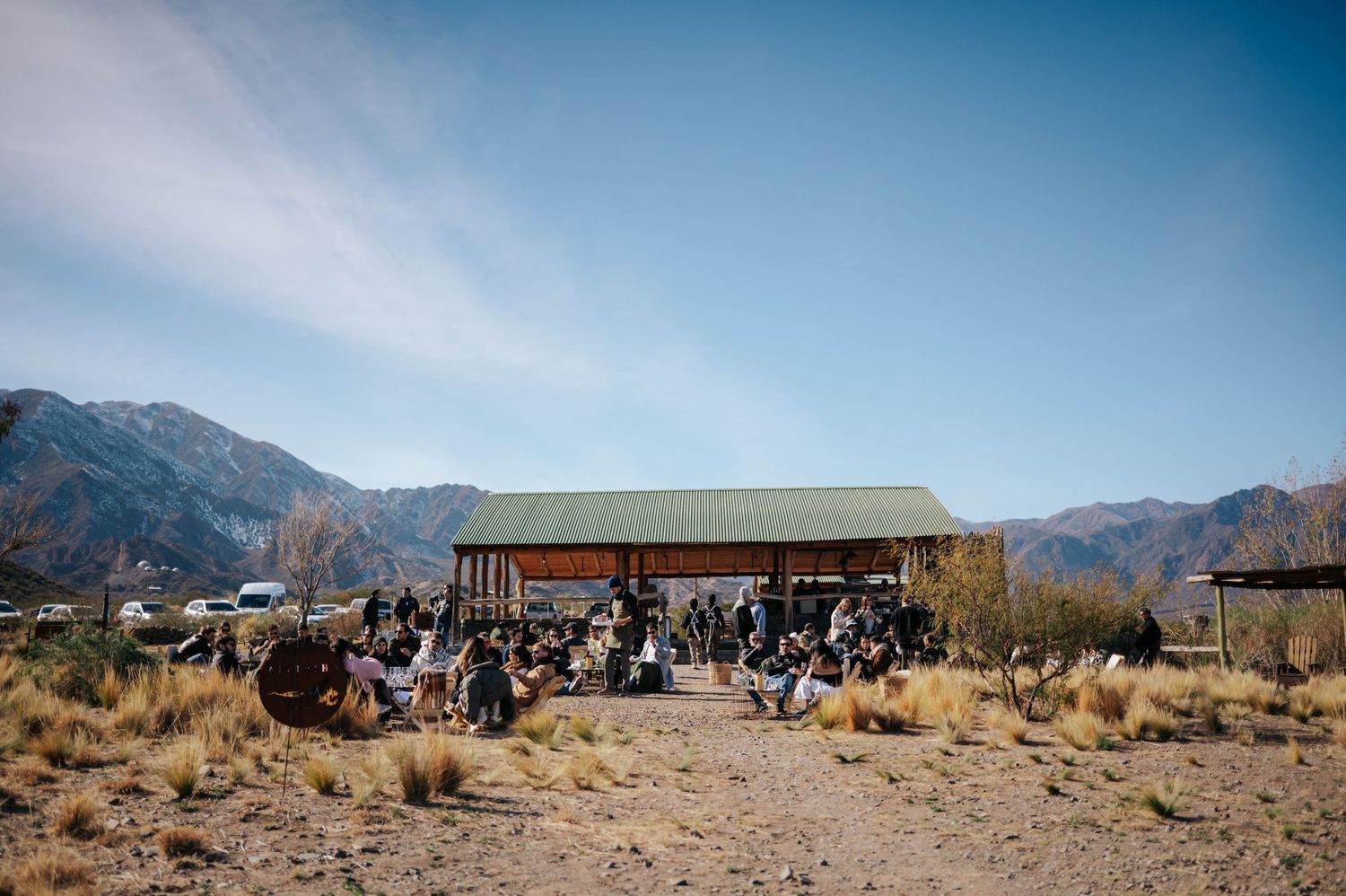 Crowd gathered at rustic open-air pavilion in arid mountain landscape under blue sky.