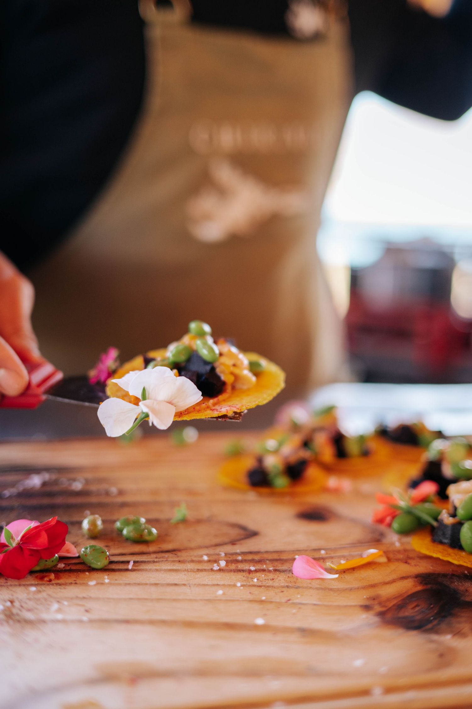 Chef plating gourmet dish with edible flowers on wooden cutting board at outdoor culinary event.