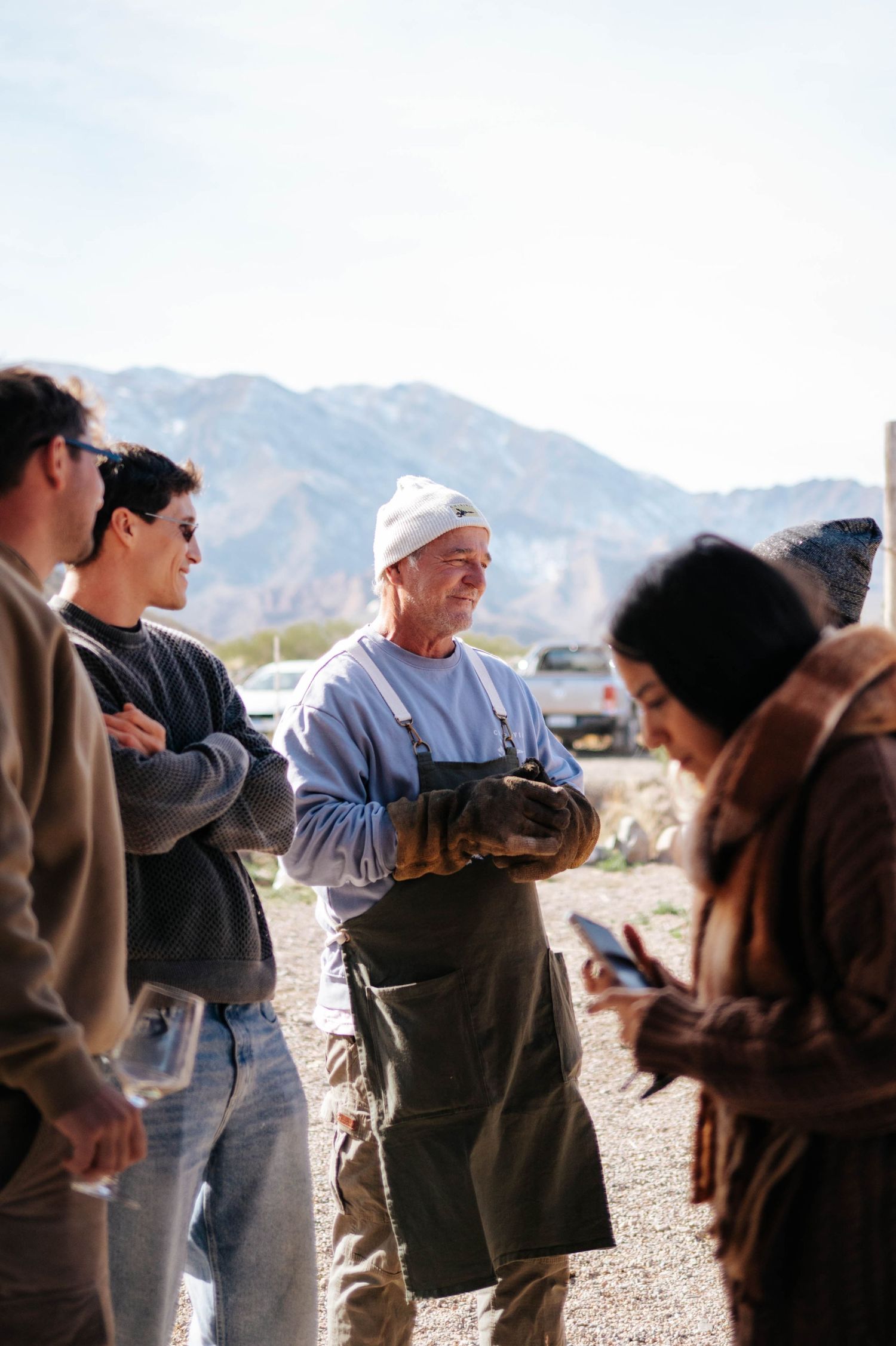 Chef in apron and white hat socializes with guests outdoors near mountain landscape.