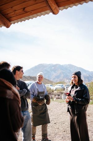 Group of people chatting with a chef in apron under rustic wooden shelter with mountain views.