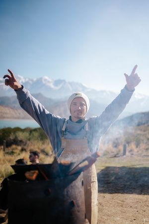 Excited chef raises arms triumphantly over open fire grill with mountain lake in background.