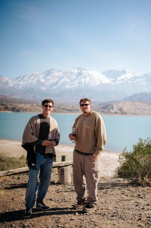 Two men smiling with drinks in front of turquoise lake and snow-capped mountain range.