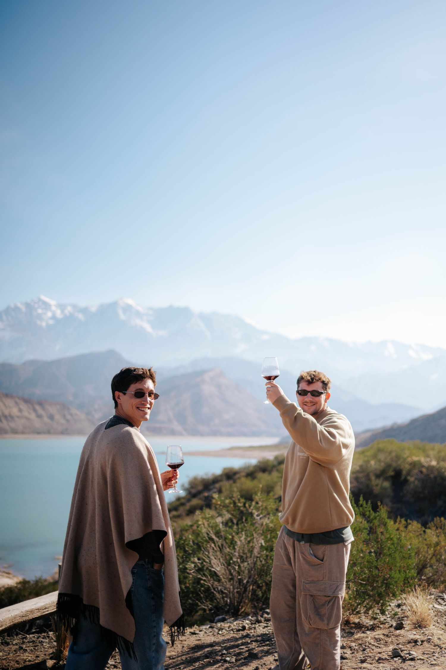 Two men toasting wine glasses with a scenic mountain lake backdrop on a sunny day.